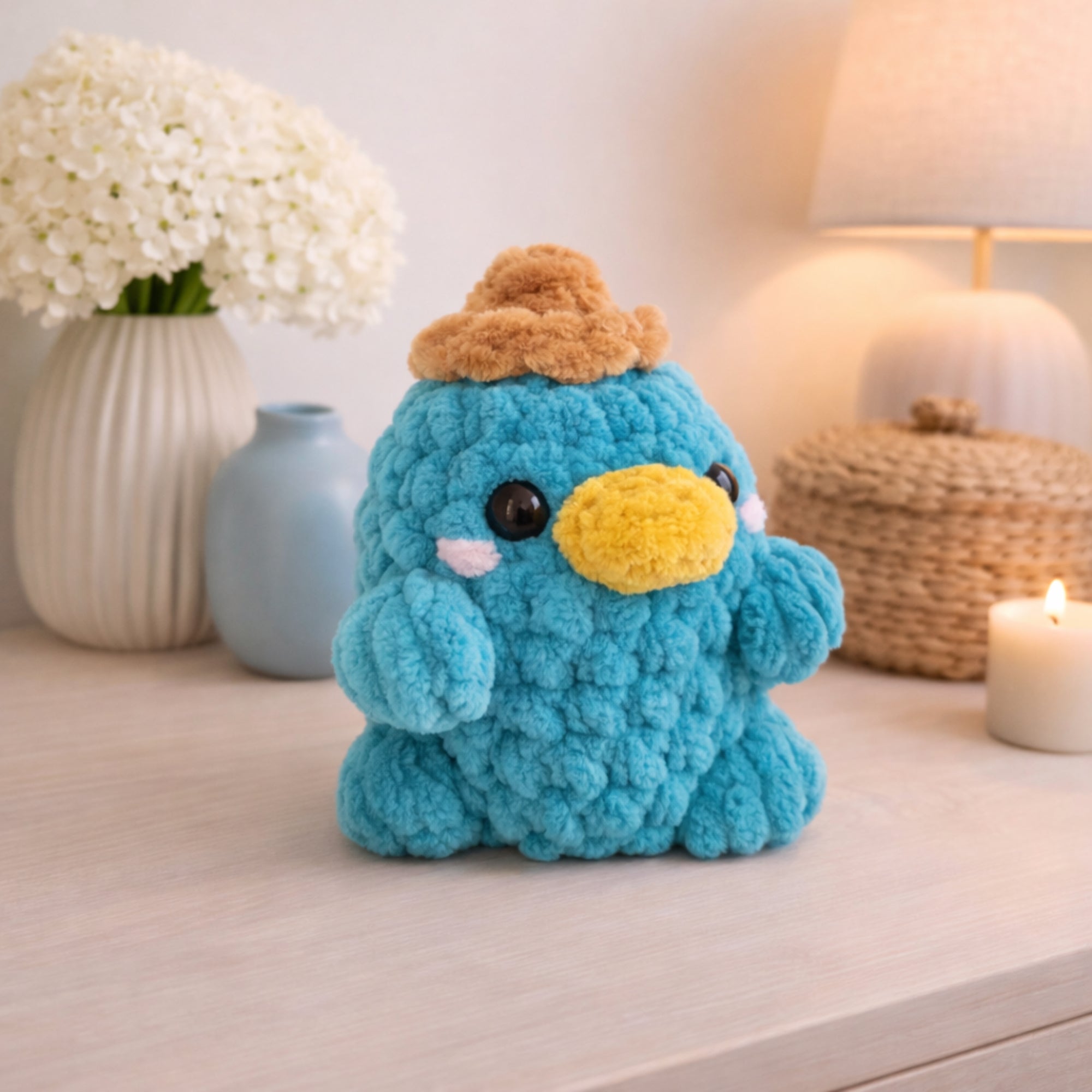 Handmade crochet platypus plush displayed as cozy home decor on a table with flowers and lamp.