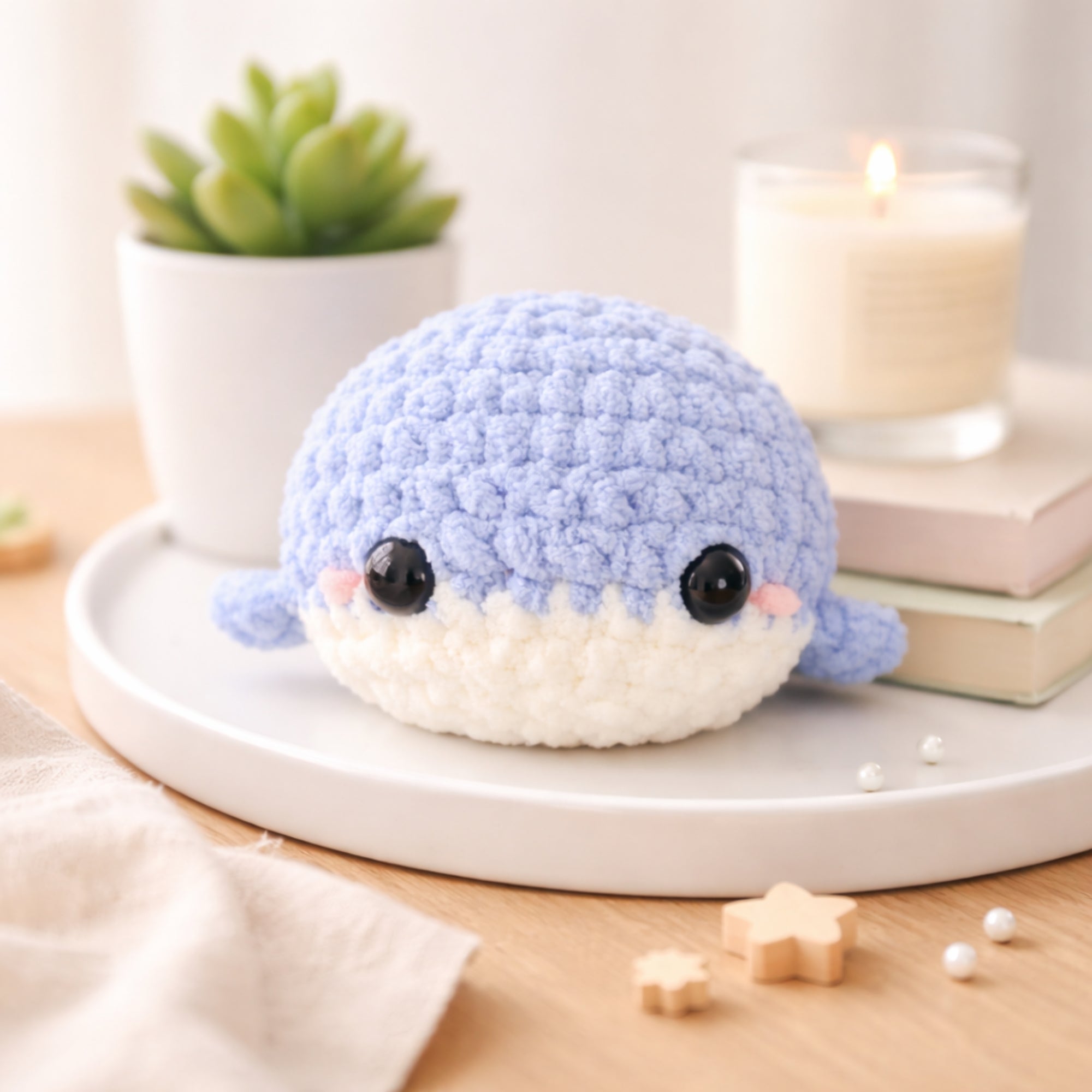 Handmade crochet baby whale amigurumi plush in blue chenille yarn displayed as cute desk decor with plant and candle